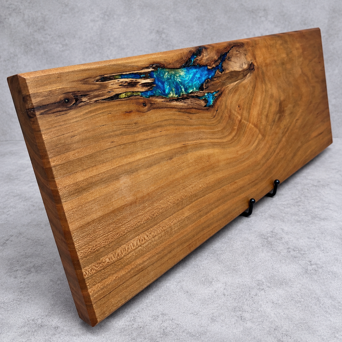 Handcrafted Hidden Lagoon wood and epoxy art serving board featuring warm honey-brown wood with sweeping natural grain and a striking turquoise, sapphire blue, and green epoxy-filled opening near the upper edge. Handmade by Trees To Home in Pennsylvania.