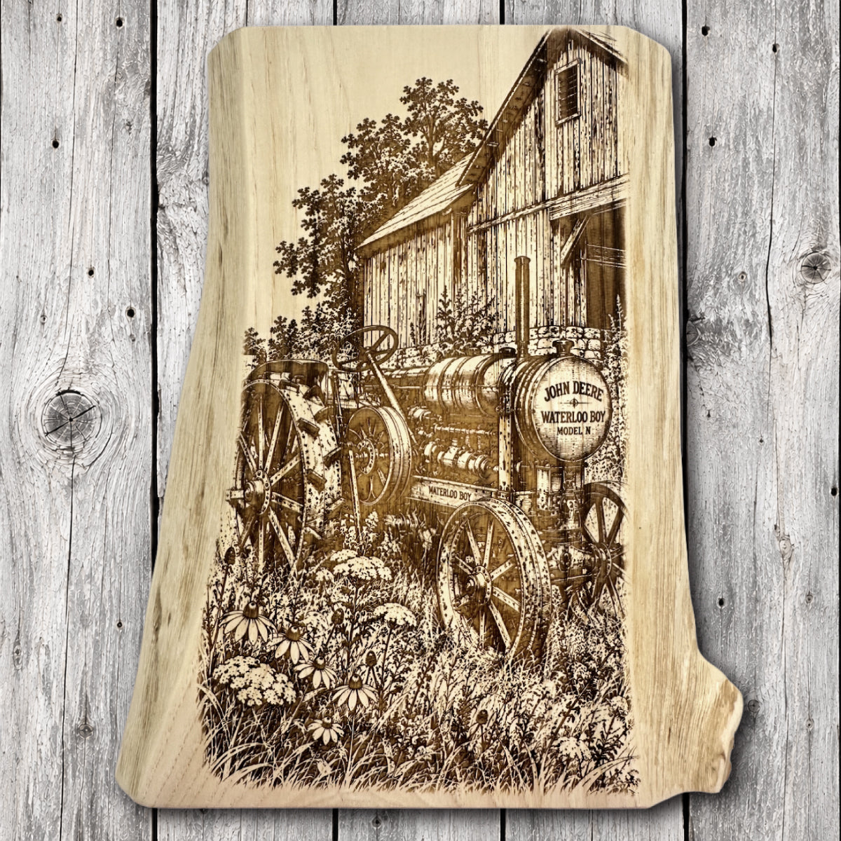 Live edge hardwood wall art featuring a detailed laser engraving of an antique John Deere Waterloo Boy tractor with steel wheels beside a rustic wooden barn surrounded by wildflowers and tall grasses.
